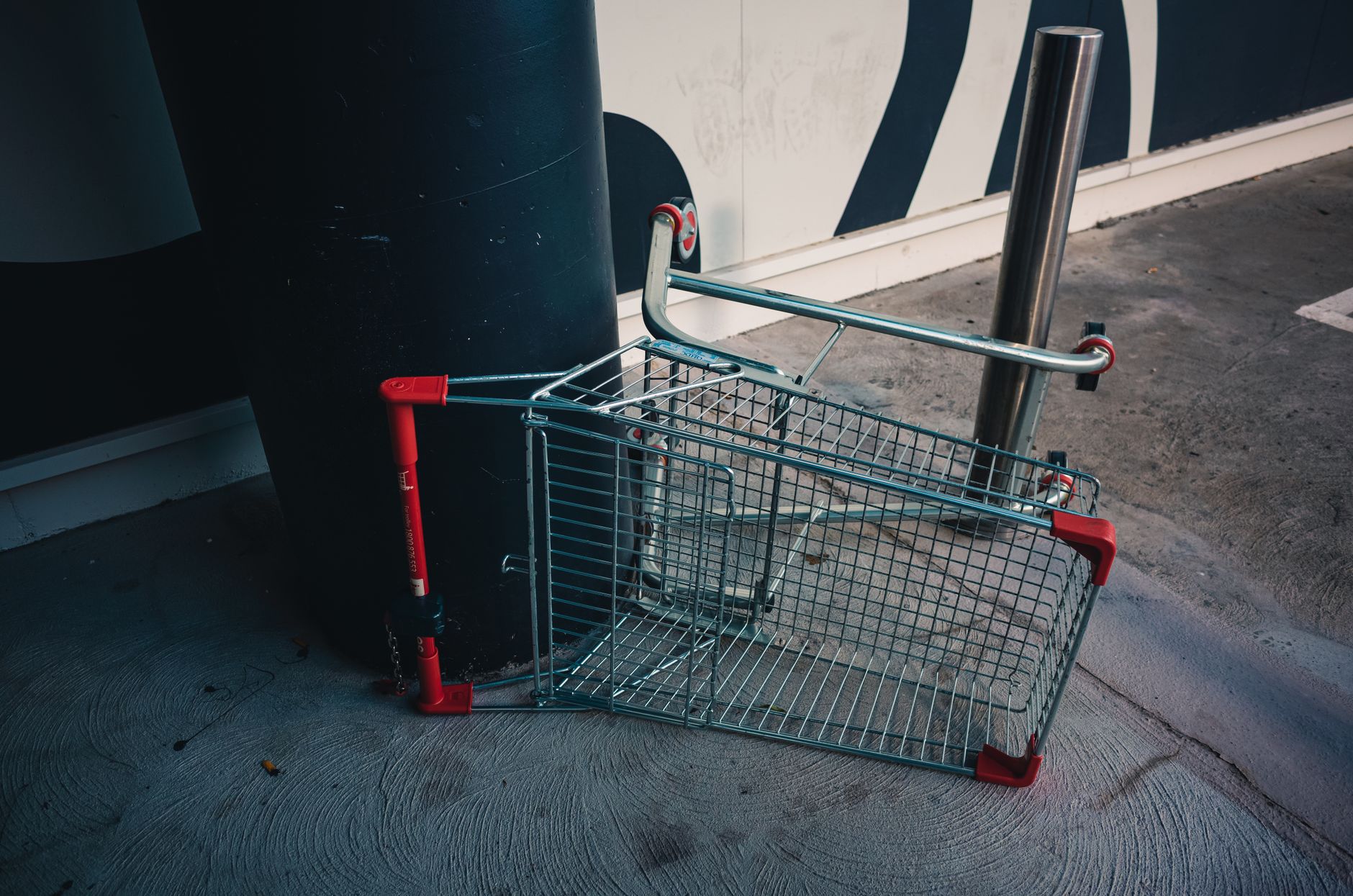 A metal shopping cart lies upside down against a building wall outdoors, indicating neglect or vandalism.