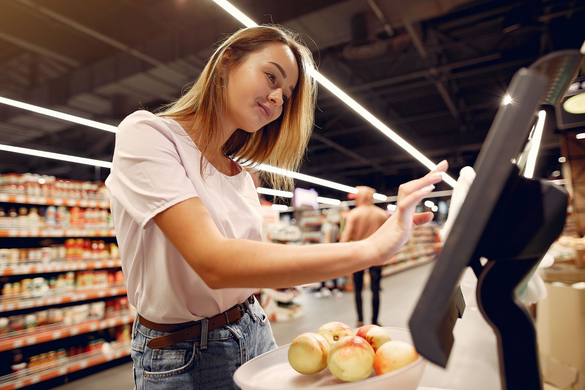 Side view of young woman in trendy clothes weighing peaches on scales while  shopping in supermarket during purchase food