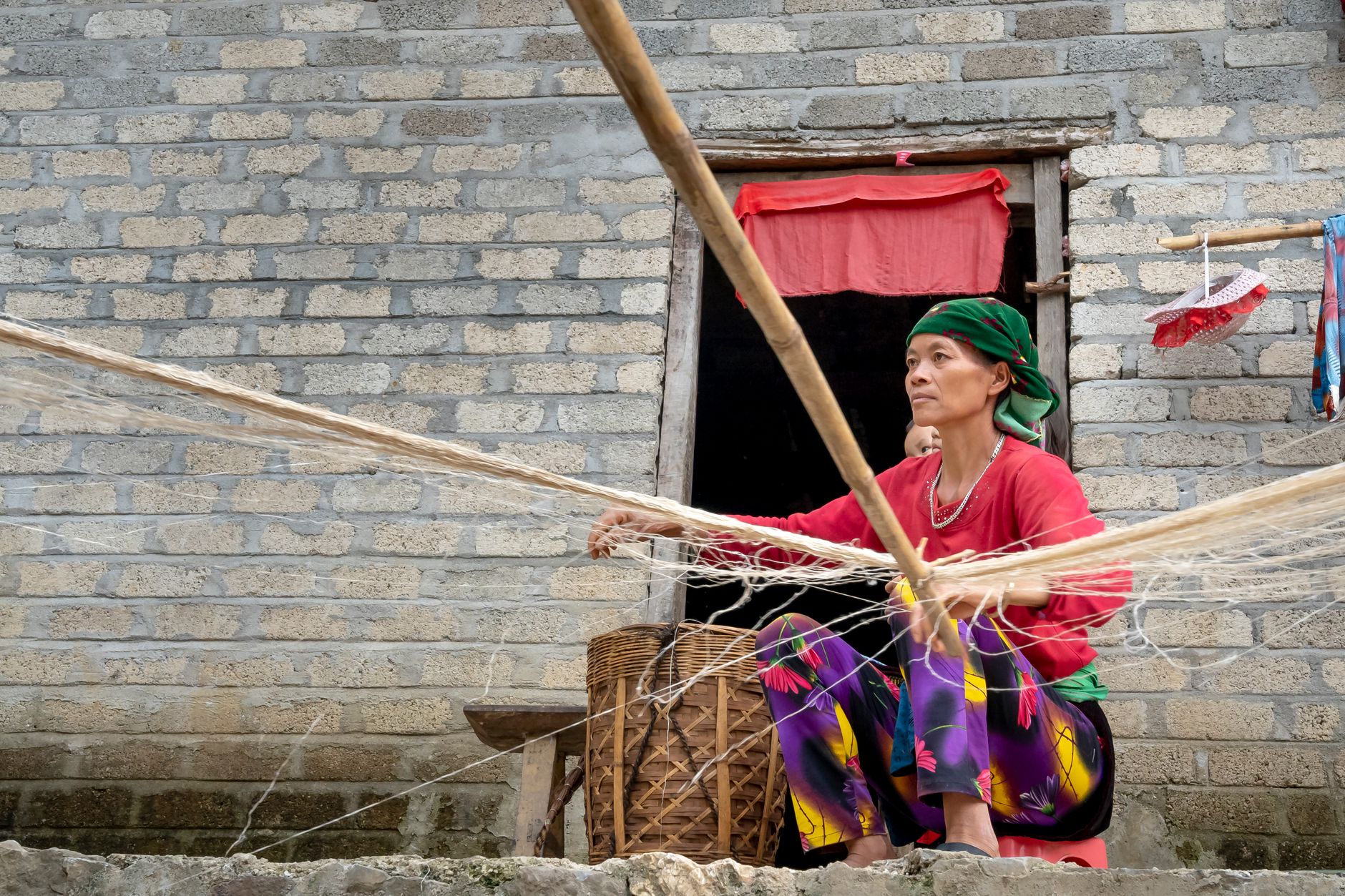 Woman weaving a fishing net outside a brick house, showcasing traditional craftsmanship.