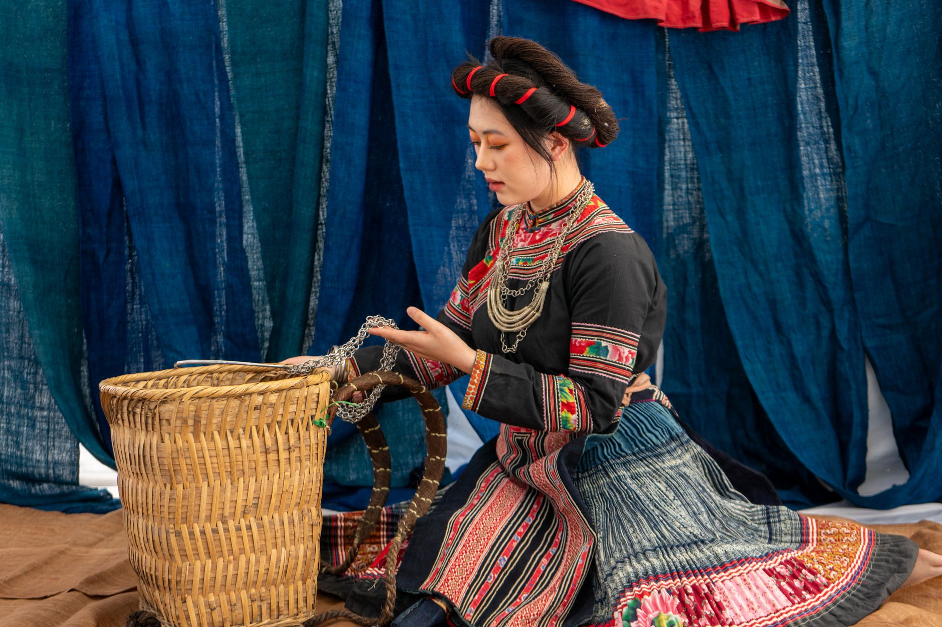 A woman in traditional attire weaving a basket in an indoor setting with blue drapes.