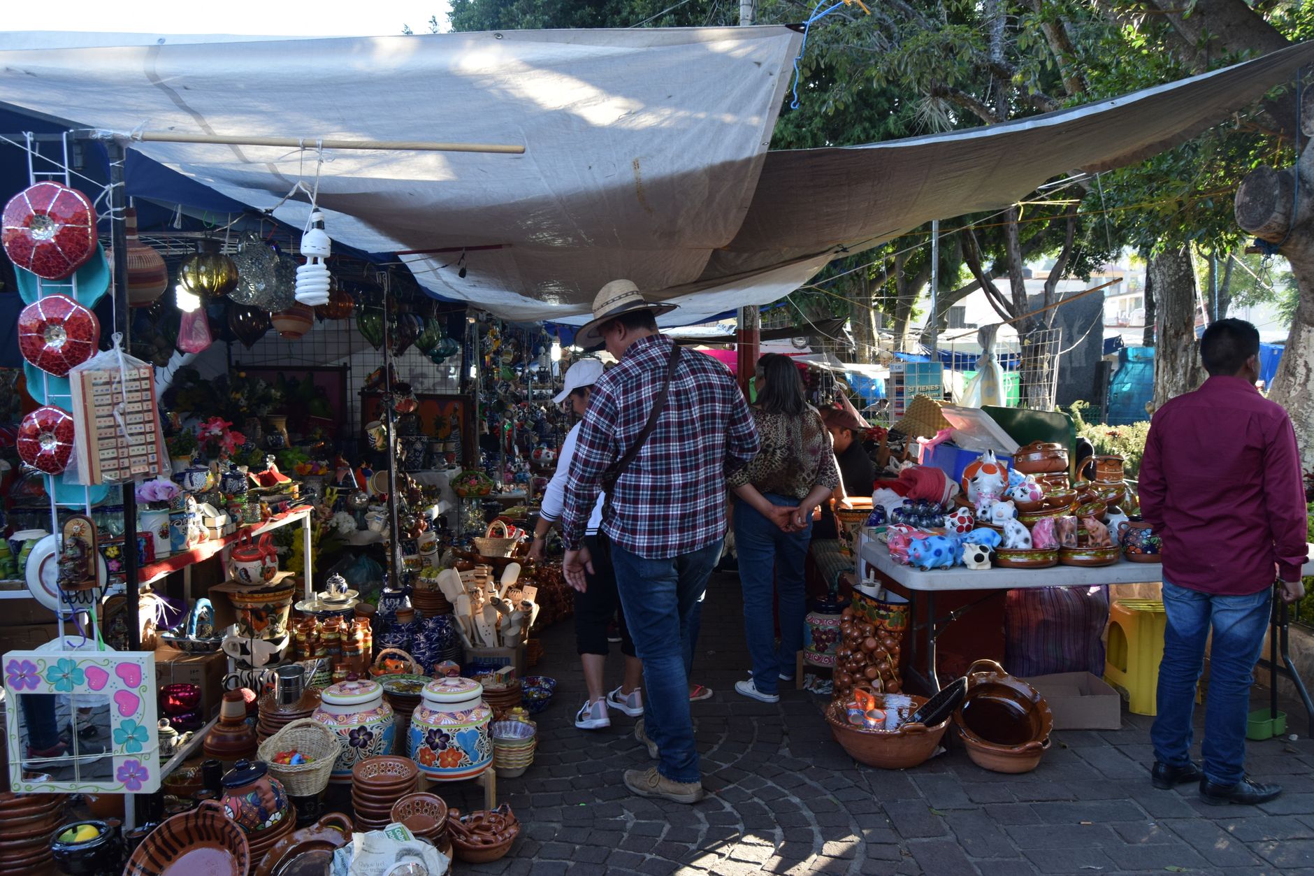 Explore a bustling outdoor market filled with pottery and vibrant souvenirs under the shade of trees.