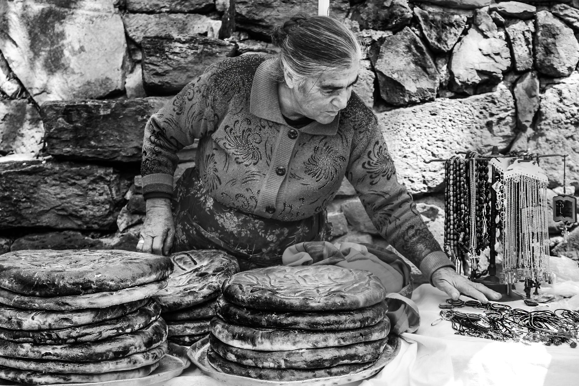 An elderly woman at a market stall sells traditional flatbread and handmade jewelry, showcasing cultural heritage.