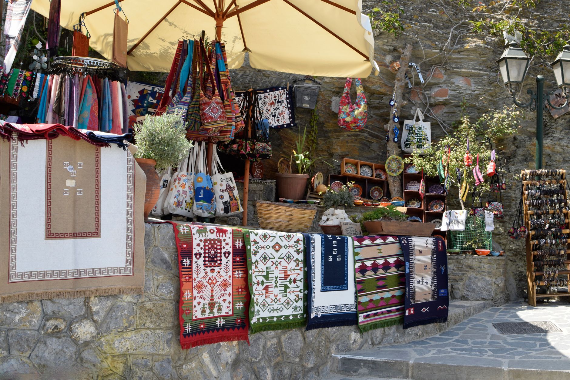 Outdoor market stall featuring vibrant textiles, rugs, and handicrafts in a sunny setting.