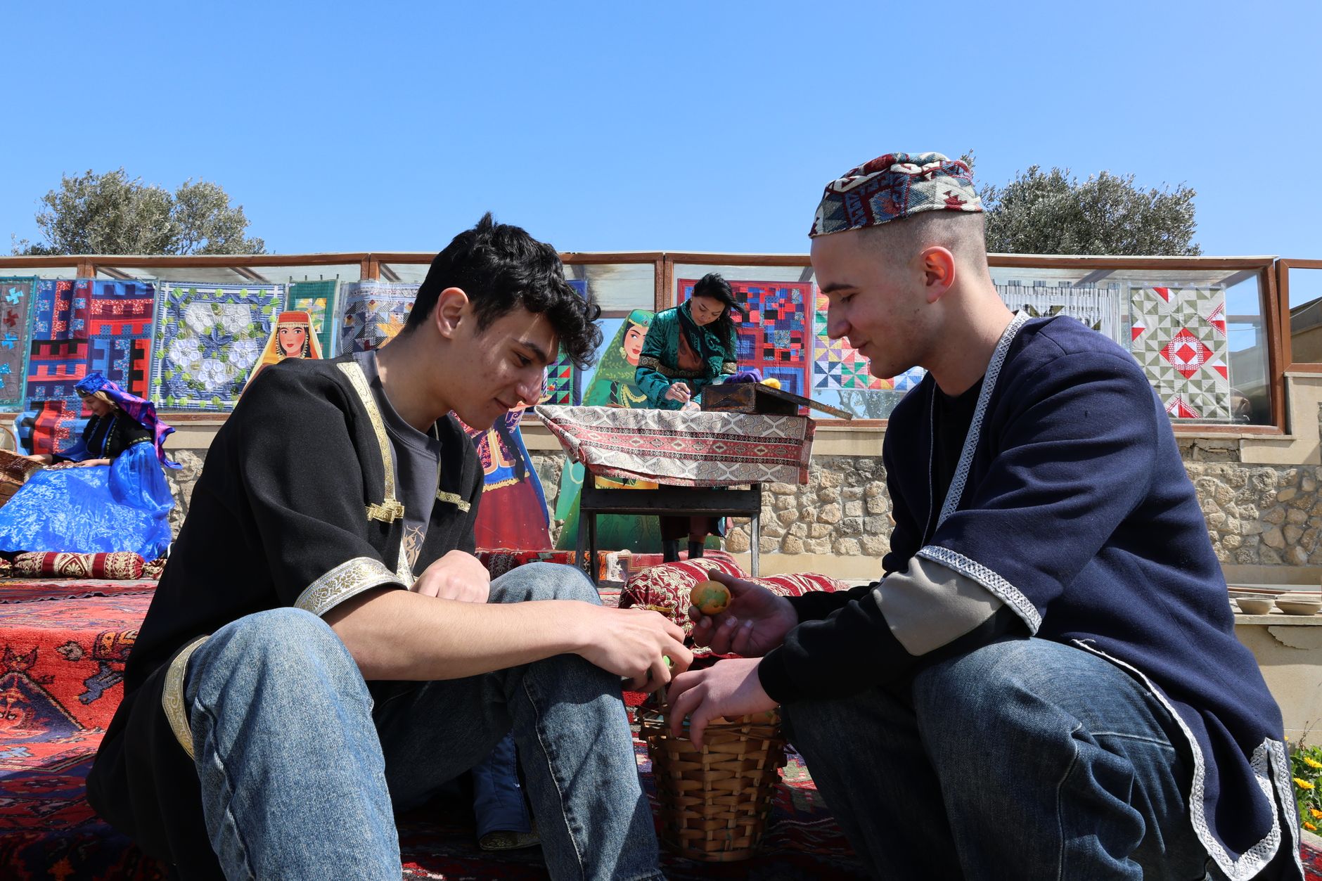 Two young adults in traditional attire engaged at an outdoor bazaar, vibrant textiles in view.