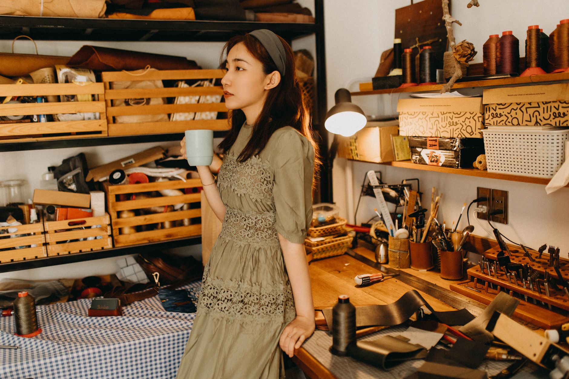 A woman sips coffee in a cozy workshop filled with tools and materials.