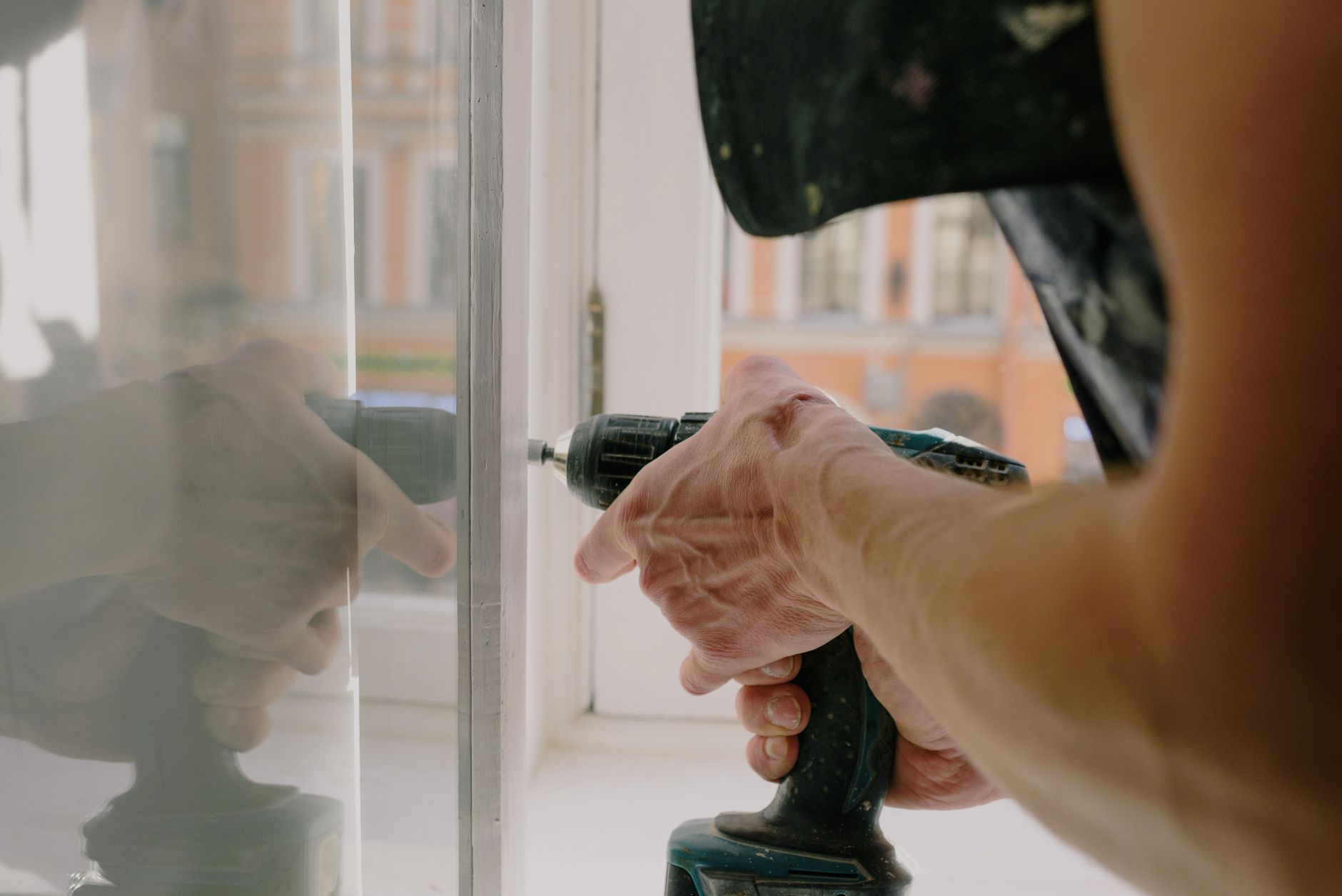 Unrecognizable professional male worker assembling white window with cordless screwdriver with drill near windowsill with reflection on glas