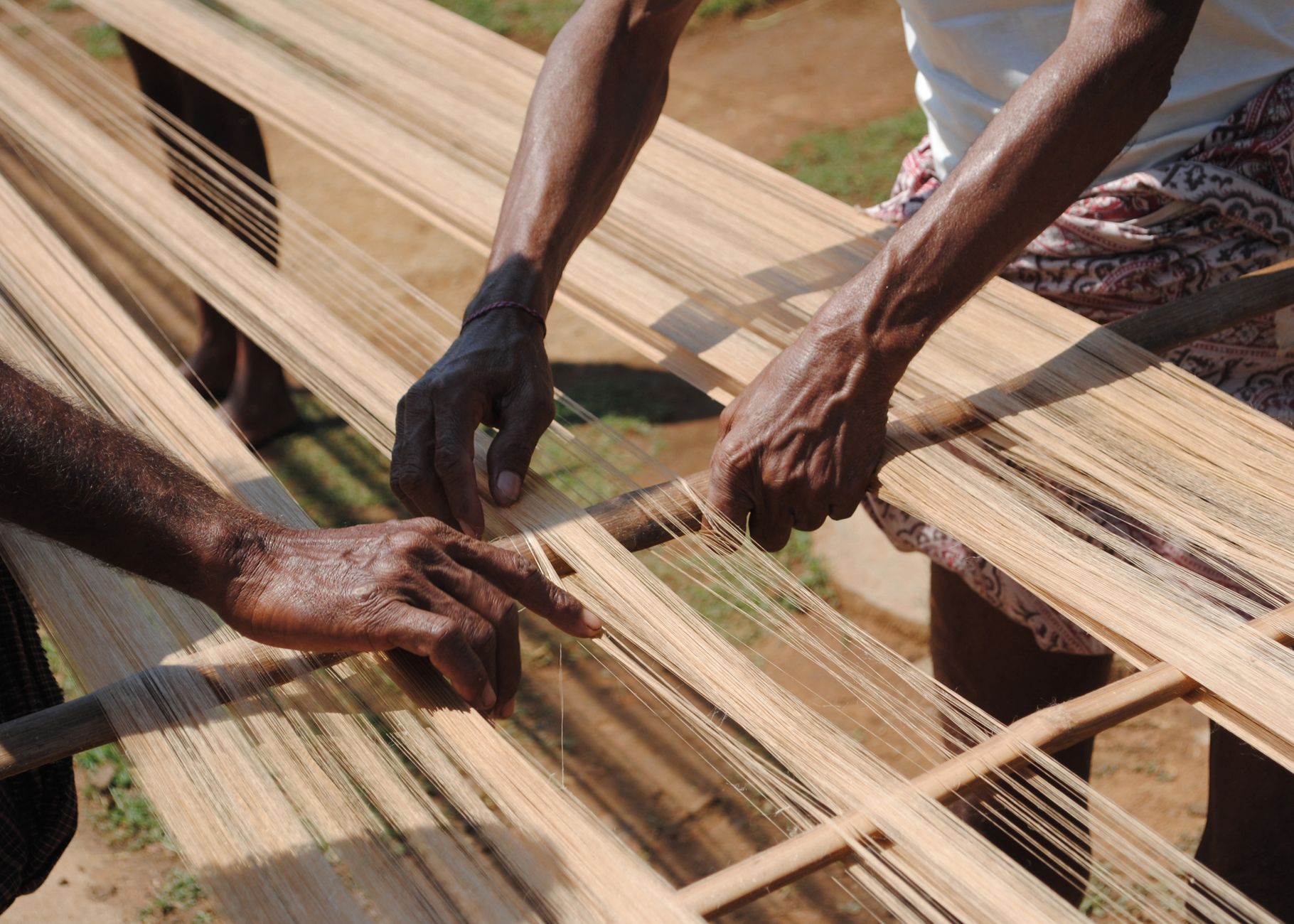 Artisans expertly weave threads on a traditional loom outdoors, showcasing skilled craftsmanship.