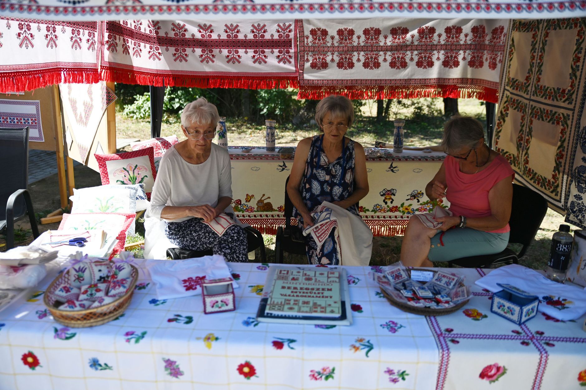 Three women showcasing handmade embroidery at a vibrant outdoor market stall.