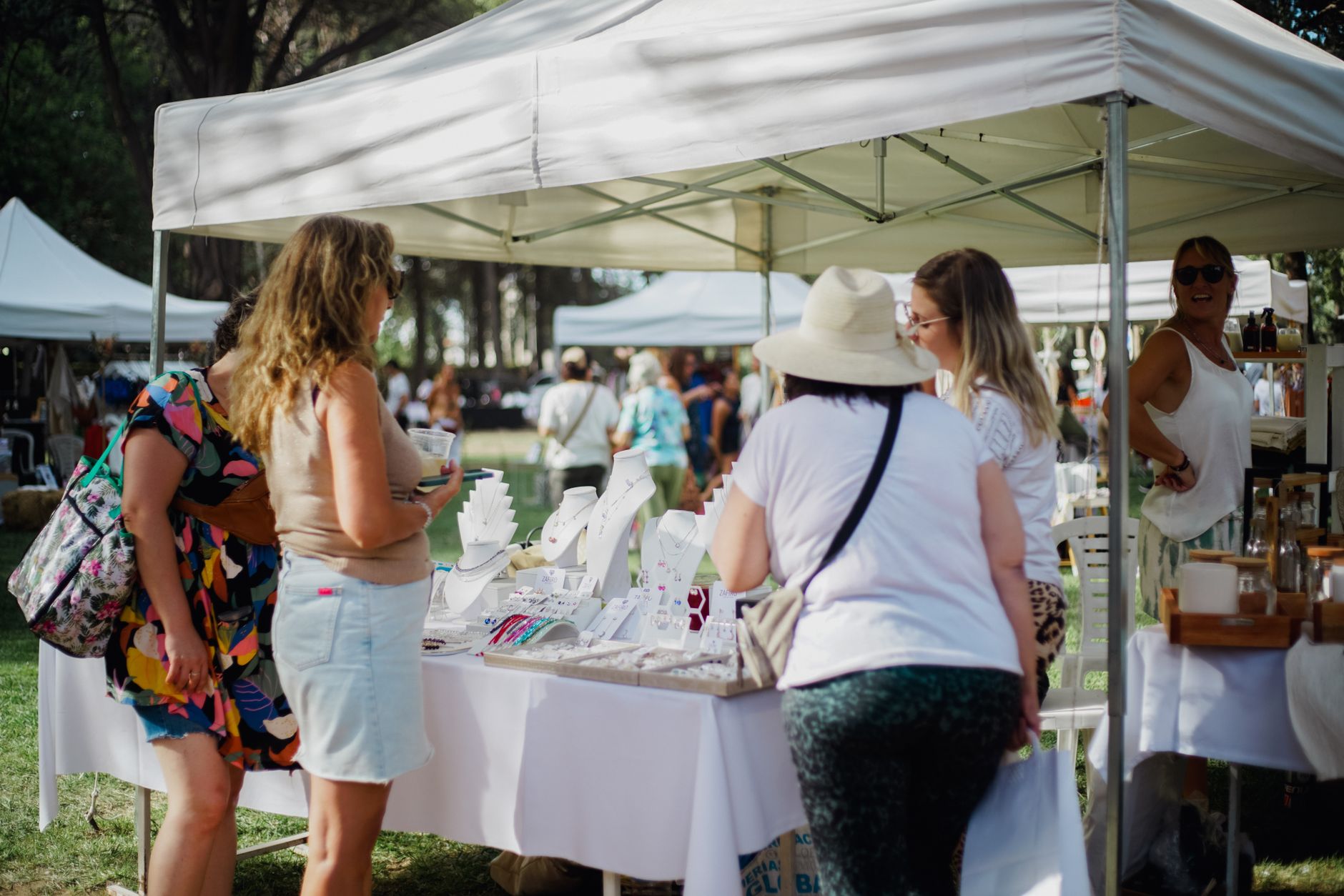 Visitors browse jewelry stalls at a bustling outdoor market on a sunny day.