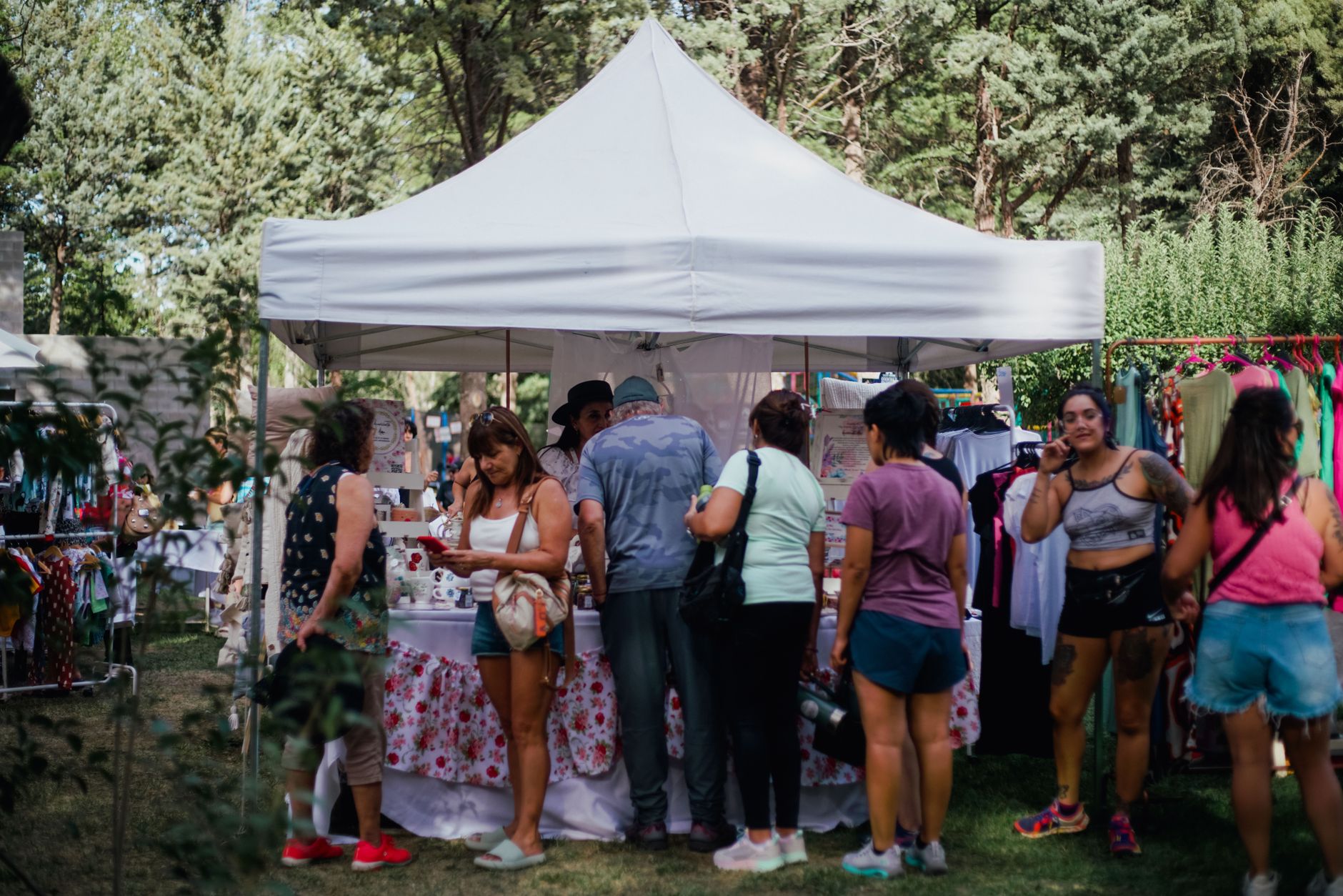 Visitors browse goods at an outdoor market stand surrounded by trees and vibrant displays.