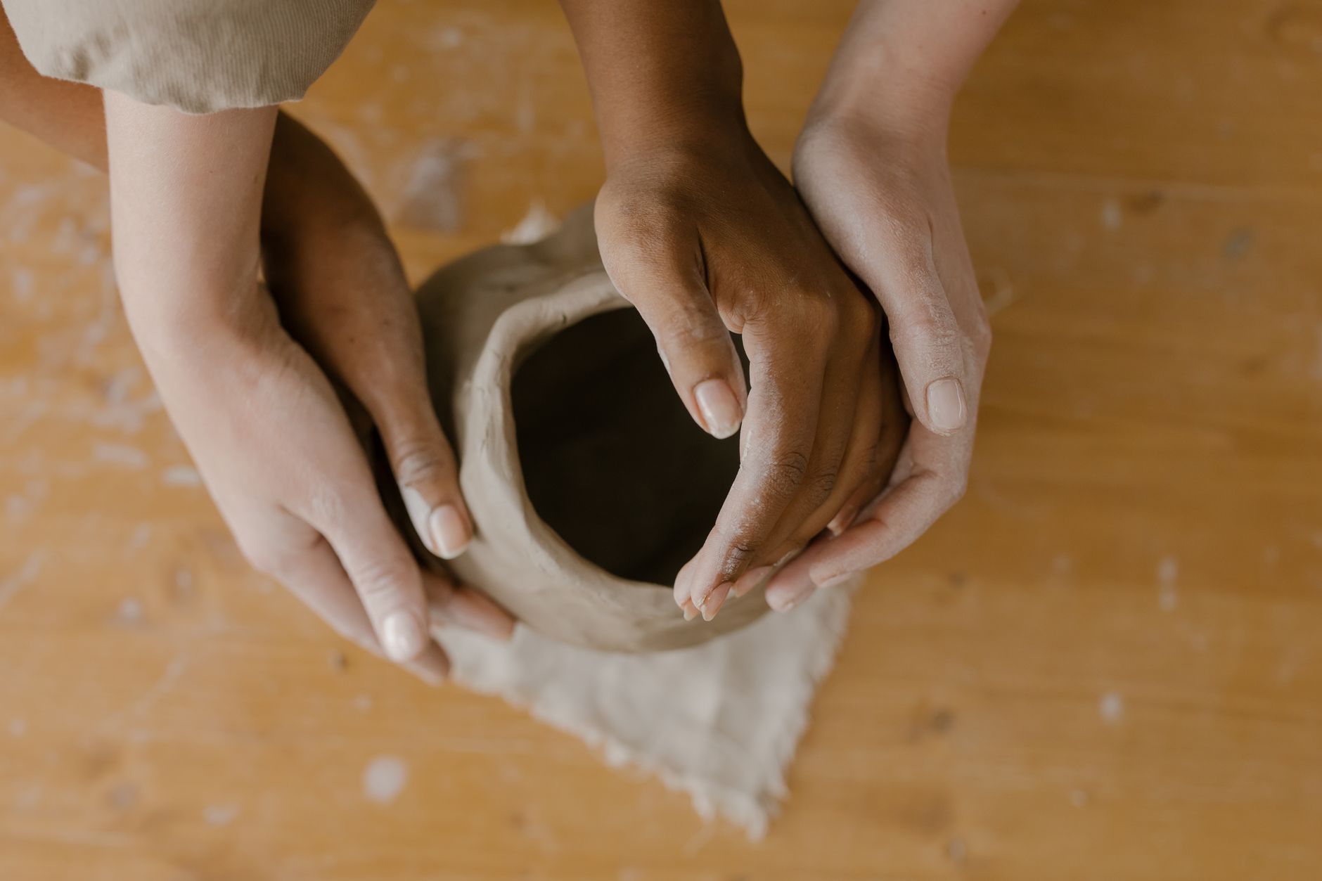 Close-up of diverse hands shaping clay pottery on a wooden table, symbolizing teamwork.