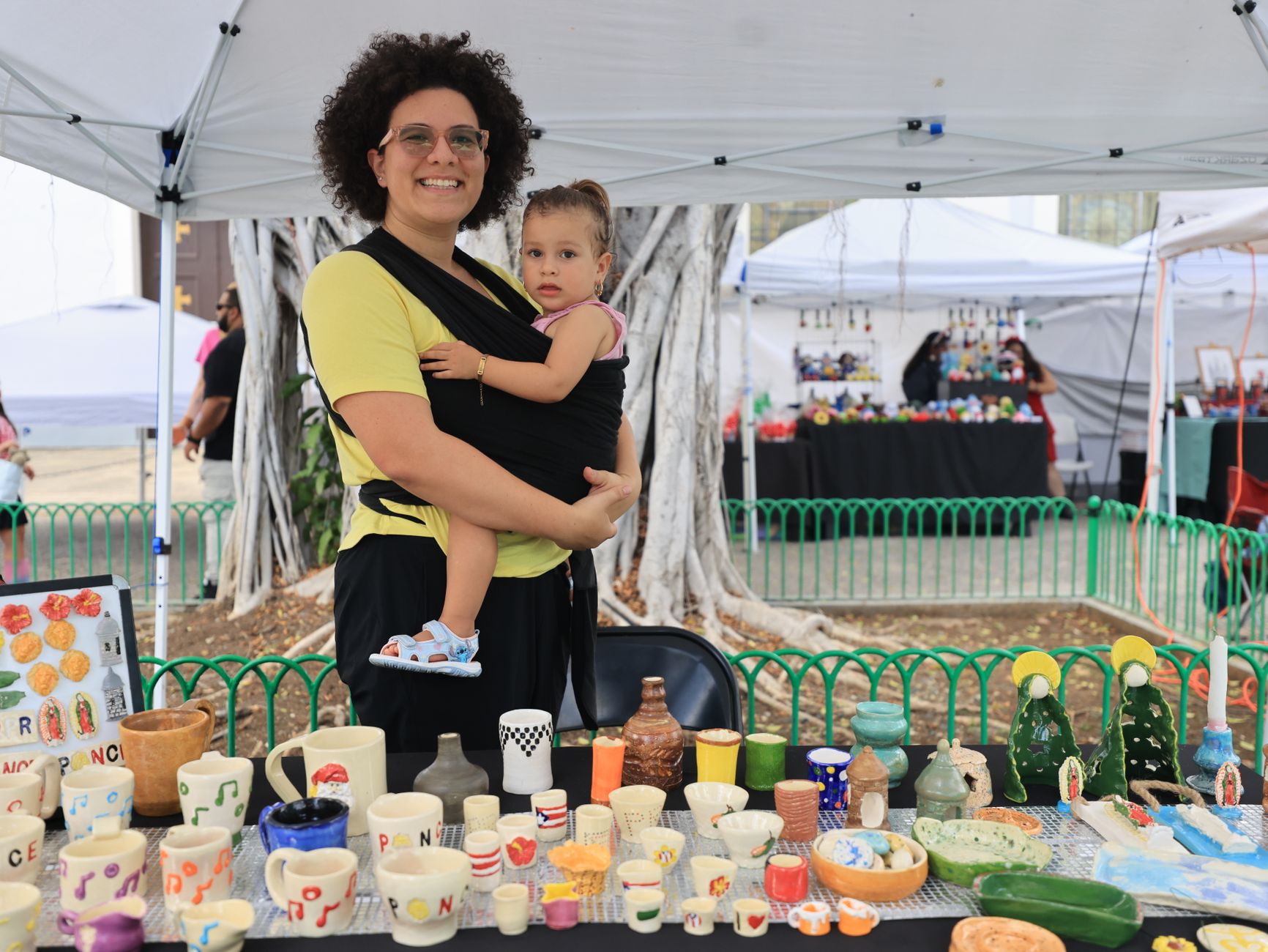 A mother holding her child at an outdoor pottery market stall showcasing colorful handmade ceramics.