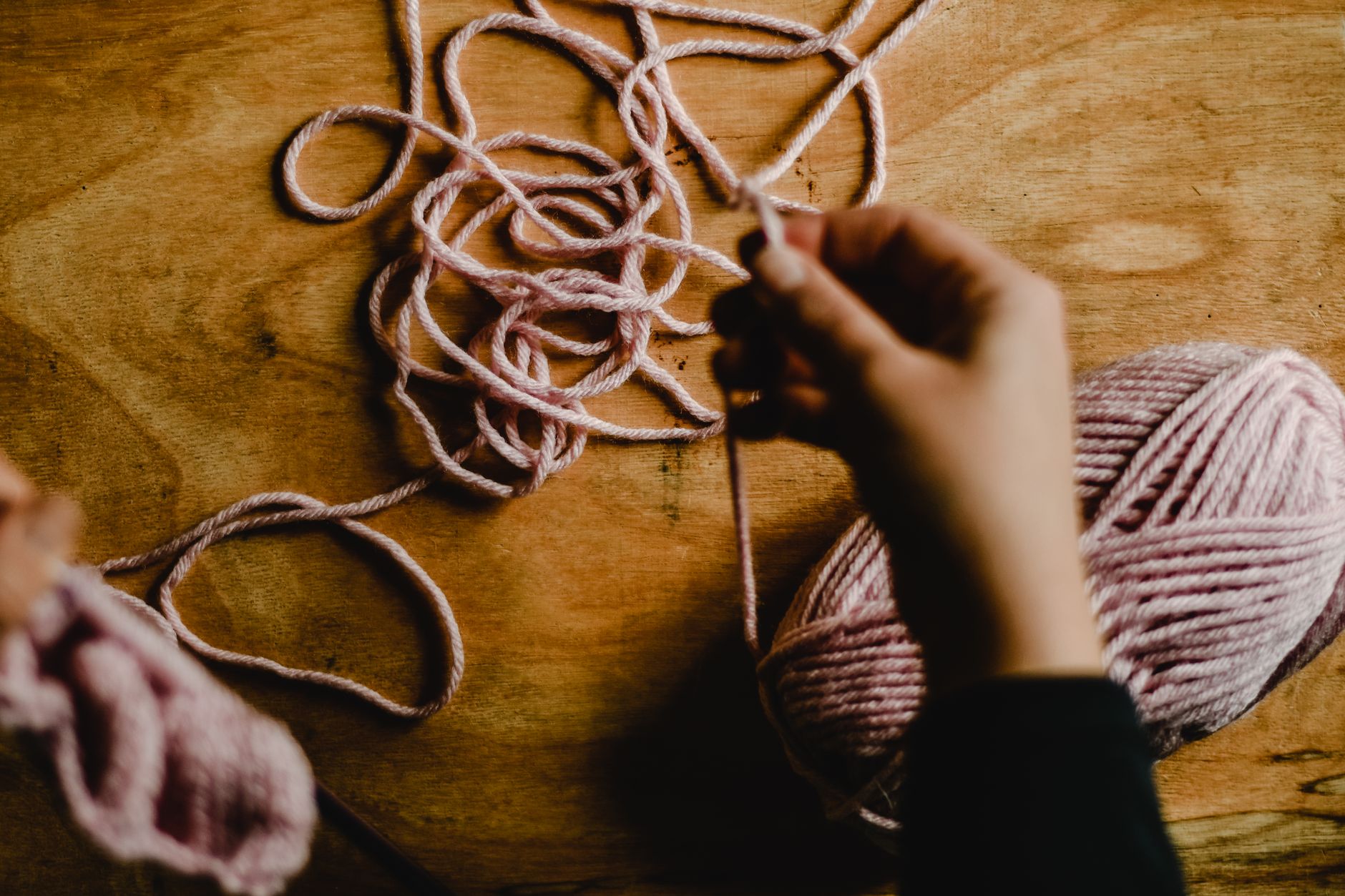 A detailed view of a person knitting with soft pink yarn on a wooden table, showcasing craftsmanship.