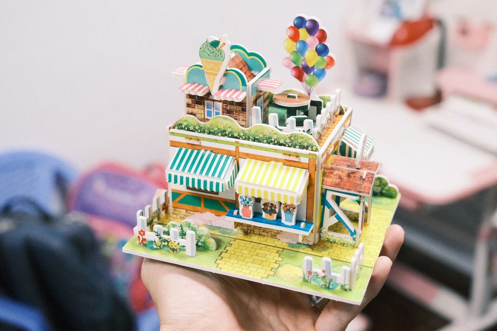 Whimsical miniature house model with vibrant balloons and pastel colors, held in hand.