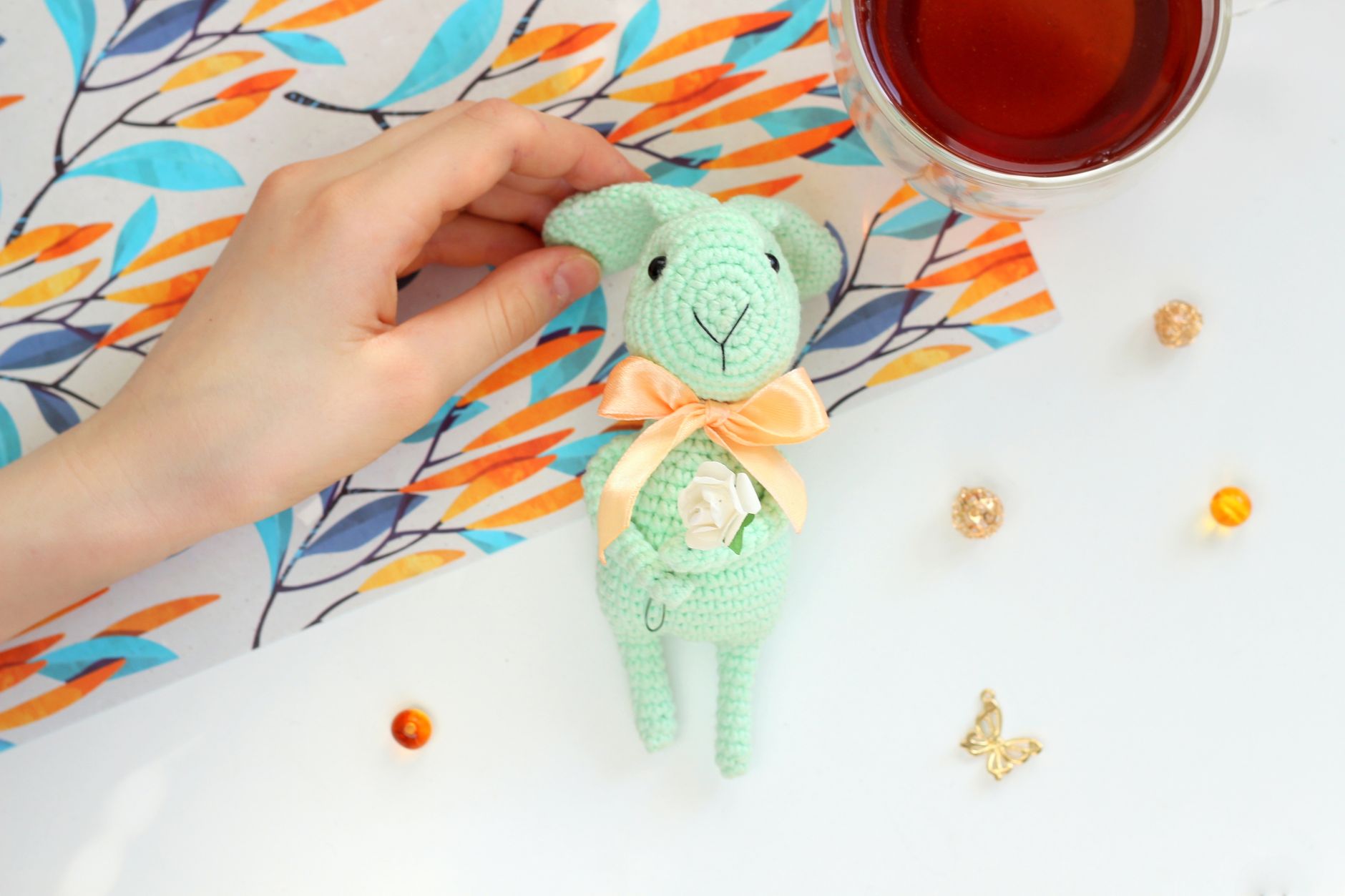 Close-up of a hand holding a crocheted bunny toy next to a cup of tea on a decorated table.