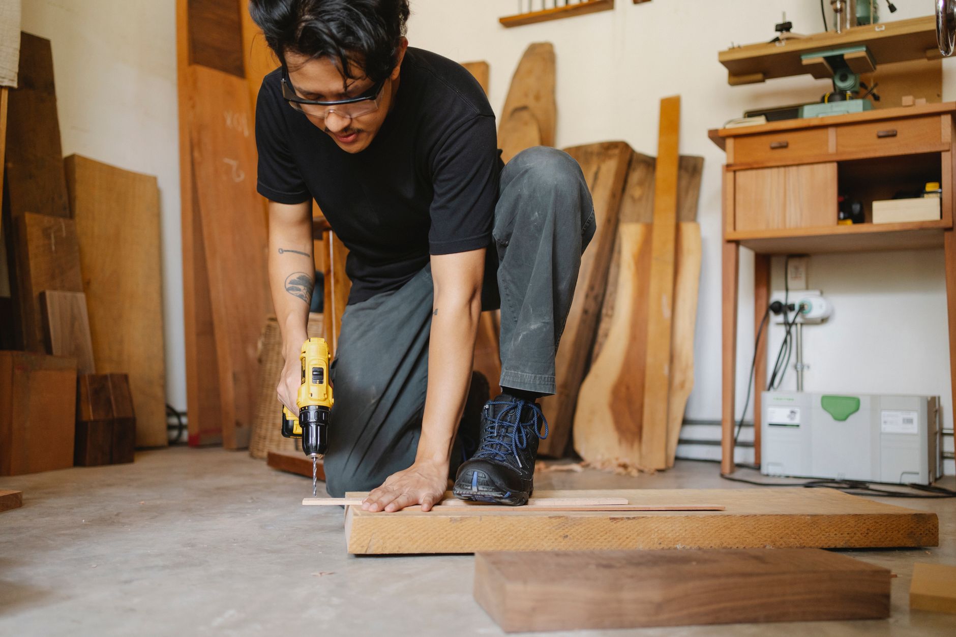 Crop ethnic male in safety glasses leaning on timber plank and drilling with screwdriver in workshop